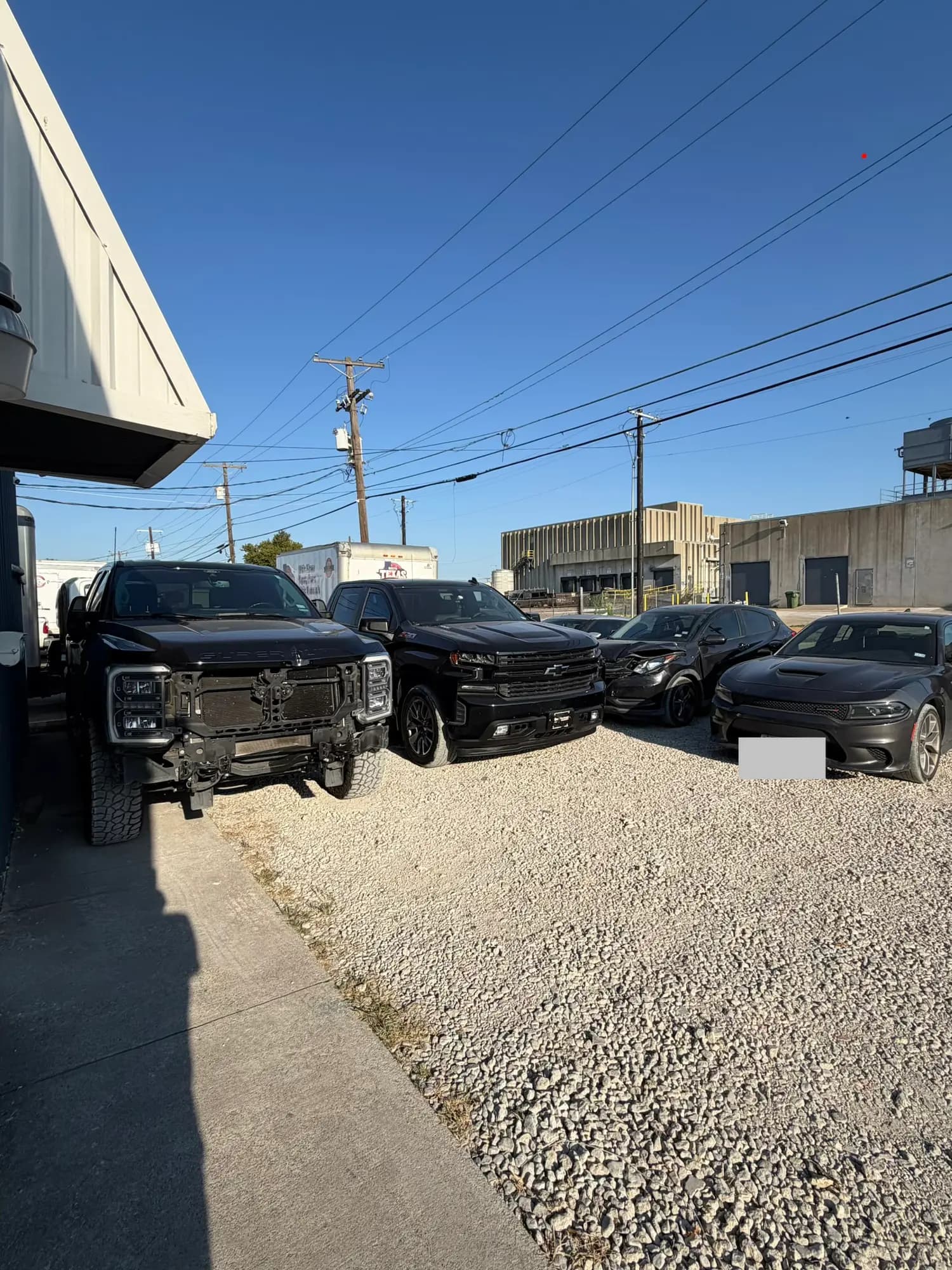 Several black vehicles, including a truck with a missing front bumper, parked on gravel outside an auto body shop